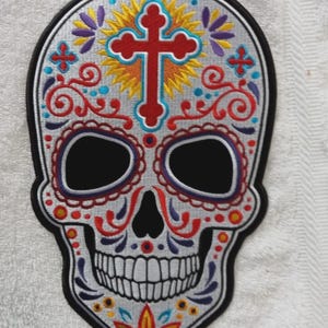 May include: An embroidered sugar skull patch featuring a central red cross surrounded by a yellow sunburst. The skull is decorated with intricate red, purple, blue, and orange floral and scroll patterns on a light gray background with a black border.
