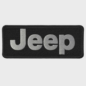 May include: A rectangular embroidered patch featuring a black background with the word Jeep stitched in light gray thread. The patch has a thick, black stitched border surrounding the entire perimeter of the rectangular shape.
