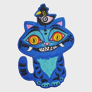 May include: An embroidered patch featuring a blue cat with large yellow eyes, a wide grin with visible fangs, and black stripes. A small black bird with a yellow beak and a white eye sits on top of the cat's head. The design uses blue, black, yellow, pink, and white thread.