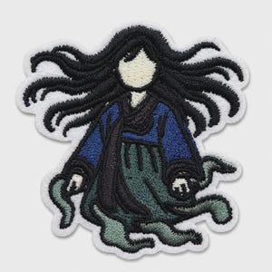 May include: An embroidered patch featuring a stylized figure with long, flowing black hair, a cream-colored face, a dark blue long-sleeved top, and a dark green skirt. The design has a white border and depicts the figure with outstretched arms.