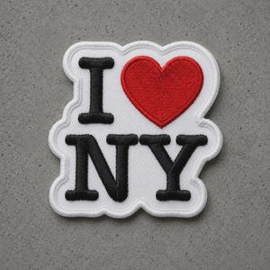 May include: A white embroidered patch featuring the black text I and NY with a red heart symbol in between. The patch has a scalloped white border and a textured fabric finish, designed to be attached to clothing or accessories as a decorative element.