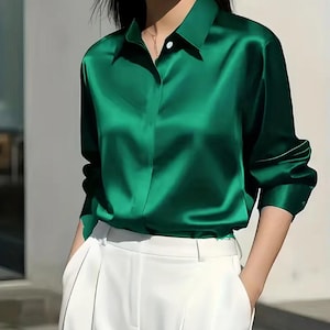May include: A long-sleeved, emerald green satin blouse with a classic collar and buttoned cuffs. The blouse is paired with white trousers. The fabric has a glossy sheen, reflecting light. The overall style is elegant and sophisticated.