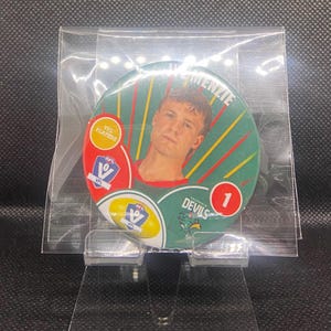 May include: A circular sports trading card featuring a portrait of a young man, with the name "W. Mckenzie" above his image. The card has a green, red, and yellow design with the text "Tasmania Devils" and the number "1". The card is in a clear plastic sleeve.