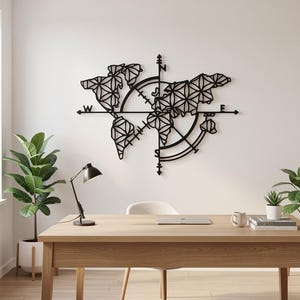 Op de afbeelding: Zwarte metalen wereldkaart wandkunst met een kompasontwerp. De kaart heeft geometrische vormen en is gemonteerd op een witte muur boven een houten bureau. Een zwarte bureaulamp en planten zijn ook zichtbaar.