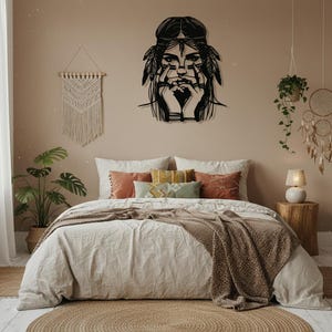 Puede incluir: Una obra de arte mural de metal negro que representa a una mujer nativa americana con plumas en el pelo, manos cerca de la cara. El arte cuelga sobre una cama con ropa de cama blanca, almohadas decorativas y una manta tejida marrón.