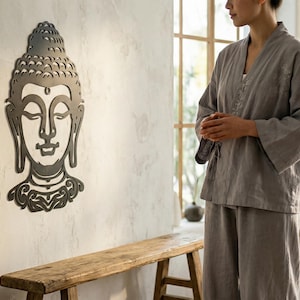 Op de afbeelding: Een metalen Buddha-hoofd wandsculptuur met gedetailleerde kenmerken is bevestigd aan een getextureerde muur. Een persoon in een grijze linnen outfit staat in de buurt. De kamer heeft een minimalistische, zen-achtige esthetiek.