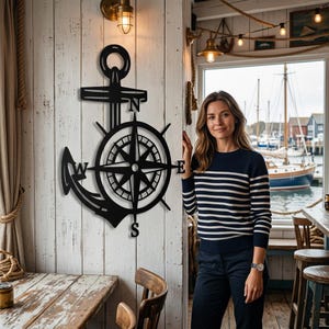 Op de afbeelding: Een zwart metalen wanddecoratie bestaande uit een scheepsanker gecombineerd met een kompasroos. Het kompas bevat de windrichtingletters N, Z, O en W, die respectievelijk aan de boven-, onder-, rechter- en linkerzijde zijn geplaatst.