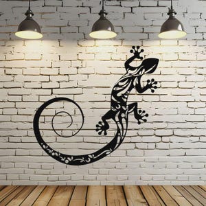 Op de afbeelding: Zwarte metalen wandkunst van een hagedisontwerp, gemonteerd op een witte bakstenen muur. De hagedis heeft een gekrulde staart. De achtergrond omvat houten vloeren en bovenverlichting.