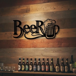 Op de afbeelding: Zwarte metalen wandkunst met de tekst "Beer" en een illustratie van een bierpul. Het bord is gemonteerd op een houten muur met verschillende bierflessen eronder. Rechts is een spiegel met lichtjes te zien.