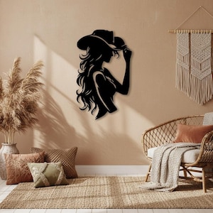 Op de afbeelding: Zwarte metalen wandkunst-silhouet van een vrouw met een cowboyhoed. Het kunstwerk toont een vrouw in profiel, met lang, golvend haar, en is gemonteerd op een beige muur. De kunst is een decoratief stuk voor een huis.