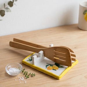 Könnte beinhalten: Eine rechteckige Löffelablage aus Keramik mit weißer Oberfläche, gelbem Rand und einem aufgemalten Zitronen- und Blattmotiv. Eine helle, hölzerne Küchenzange mit drei Schlitzen im Kopf liegt horizontal auf der integrierten weißen Halterung.