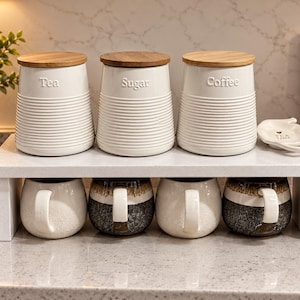 May include: Three white ceramic canisters with horizontal ribbed textures and bamboo lids labelled Tea, Sugar, and Coffee, sitting on a white shelf above four mugs. Two mugs are solid white, and two are two-toned brown and black with white handles.