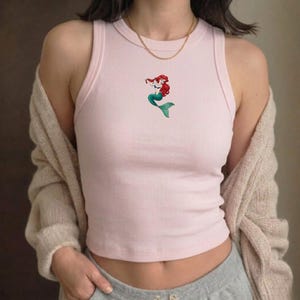 May include: A light pink ribbed tank top featuring an embroidered image of a mermaid with red hair and a green tail. The top has a round neckline and a sleeveless design. A gold-colored necklace is visible.