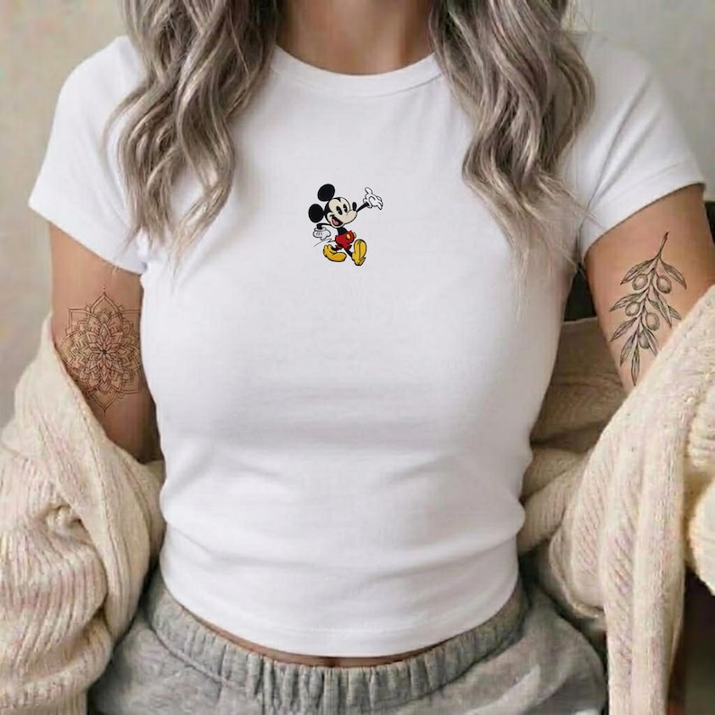 May include: White short-sleeved t-shirt featuring a small, colorful graphic of Mickey Mouse. The cartoon character is depicted in a classic pose, with black ears, a red and yellow outfit, and white gloves. The shirt is a simple, casual style.