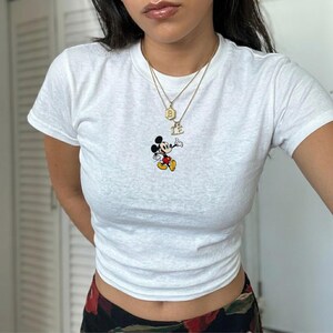 May include: White t-shirt featuring a small, colorful Mickey Mouse graphic. The shirt has a crew neck and short sleeves. A gold necklace with a pendant is visible. The shirt is a casual style.