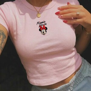 Pu&ograve; includere: Un crop top rosa chiaro con un motivo di Minnie Mouse. Il motivo include il volto di Minnie, un fiocco rosso e le parole "Minnie Mouse" in corsivo nero. Il top ha maniche corte e scollo tondo.