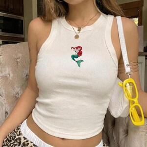 May include: Cream-colored tank top featuring a cartoon mermaid design with red hair and a green tail. The sleeveless top has a round neckline and is made of a soft, ribbed fabric. The top is paired with a white purse and yellow sunglasses.