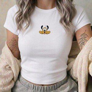 May include: A white short-sleeved t-shirt featuring a small, centered graphic of the cartoon character Pluto. The dog is yellow with black ears and a small pink tongue. The shirt is worn with a beige knit cardigan and grey sweatpants.