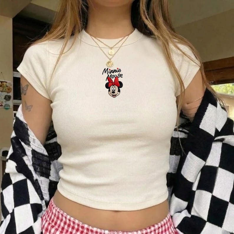 Pu&ograve; includere: T-shirt color crema a maniche corte con texture a coste. La maglietta presenta una grafica di Minnie Mouse con un fiocco rosso e la scritta "Minnie Mouse". Una giacca a scacchi bianchi e neri &egrave; drappeggiata sulle spalle.