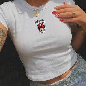 Pu&ograve; includere: Top corto bianco con grafica di Minnie Mouse. Il disegno mostra il volto di Minnie con un fiocco rosso e la scritta "Minnie Mouse". La maglietta ha girocollo e maniche corte.