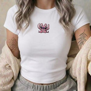 Op de afbeelding: Een wit T-shirt met korte mouwen en een ronde hals, voorzien van een kleine, centraal geplaatste afbeelding van een roze stripfiguur met grote oren en donkere ogen. Het shirt wordt gedragen met een beige gebreid vest en een grijze joggingbroek.