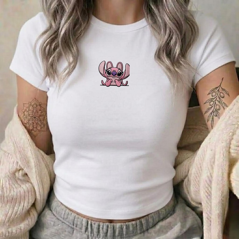 Op de afbeelding: Een wit T-shirt met korte mouwen en een ronde hals, voorzien van een kleine, centraal geplaatste afbeelding van een roze stripfiguur met grote oren en donkere ogen. Het shirt wordt gedragen met een beige gebreid vest en een grijze joggingbroek.