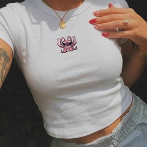 Op de afbeelding: Een wit T-shirt met korte mouwen en een ronde hals, voorzien van een kleine geborduurde afbeelding van een roze stripfiguur met grote oren en donkere ogen op de borst, gecombineerd met een gouden ketting en een gouden ring met een heldere steen.