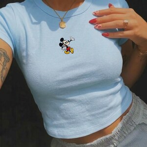 May include: A light blue crop top featuring a small Mickey Mouse graphic. The cartoon character is depicted in black, white, yellow, and red. The top has short sleeves and a round neckline, made of a soft, stretchy material.