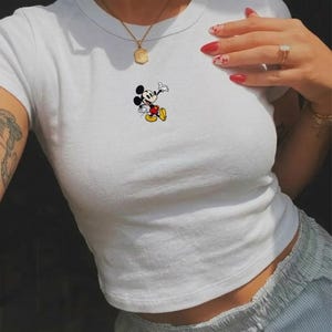 May include: White crop top featuring a small Mickey Mouse graphic. The cartoon character is depicted in his classic pose, with black ears, a red and yellow outfit, and white gloves. The shirt has a round neckline and short sleeves.