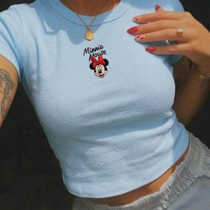 Pu&ograve; includere: Top corto azzurro con un disegno di Minnie Mouse. Il disegno presenta il volto di Minnie, un fiocco rosso e le parole "Minnie Mouse" in corsivo nero. Il top ha maniche corte e girocollo.