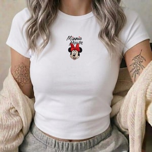 Pu&ograve; includere: T-shirt bianco con grafica di Minnie Mouse. Il design include il volto di Minnie, un fiocco rosso e la scritta "Minnie Mouse" in nero. La maglietta ha maniche corte e girocollo.