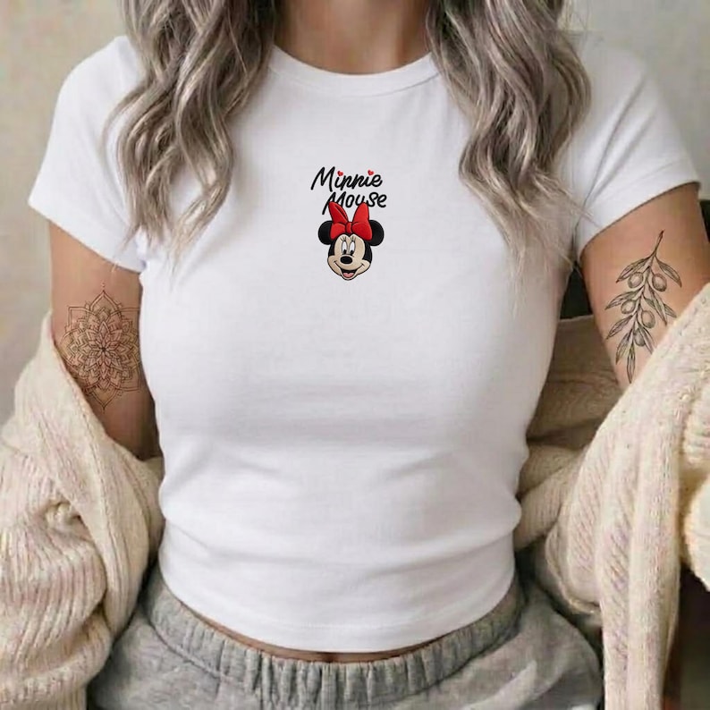 Pu&ograve; includere: T-shirt bianco con grafica di Minnie Mouse. Il design include il volto di Minnie, un fiocco rosso e la scritta "Minnie Mouse" in nero. La maglietta ha maniche corte e girocollo.