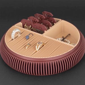 May include: A maroon and beige jewellery organiser with multiple compartments. The round organiser has a ribbed exterior and a slotted interior. Various rings, earrings, and necklaces are displayed inside the organiser.