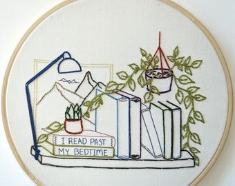 Könnte beinhalten: Stickerei in einem runden Rahmen mit einem Bücherregal mit Büchern, einer Topfpflanze, einer Hängepflanze und einer Lampe. Die Bücher tragen den Text "I read past my bedtime".