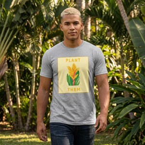 May include: A gray t-shirt with the words "PLANT THEM" in a yellow square, featuring a botanical illustration of a yellow and orange flower with green leaves. The shirt is worn by a person in a natural outdoor setting.