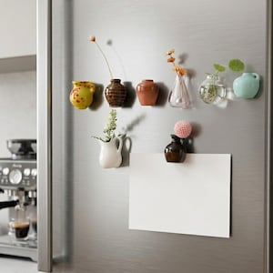 May include: A selection of miniature ceramic vases in diverse shapes and colours, affixed to a fridge. Some vases contain small flowers and greenery. A blank white paper is held by a vase.