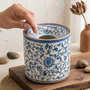 May include: A ceramic ashtray with a blue and white floral design. The cylindrical ashtray has a lid with a central opening and a decorative rim. A lit cigarette is held above the ashtray. The ashtray sits on a wooden surface.