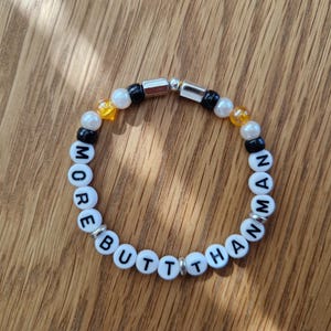Könnte beinhalten: Ein Perlenarmband mit den Worten "MORE BUTT THAN MAN" in weißen Buchstabenperlen. Das Armband hat außerdem schwarze, weiße und gelbe Perlen sowie silberfarbene Metallakzente. Das Armband wird auf einer Holzoberfläche präsentiert.
