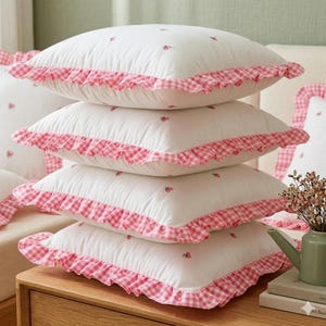 May include: Four white square cushions with pink gingham frill edging. Each cushion features small embroidered pink floral details. The cushions are stacked on a wooden surface.