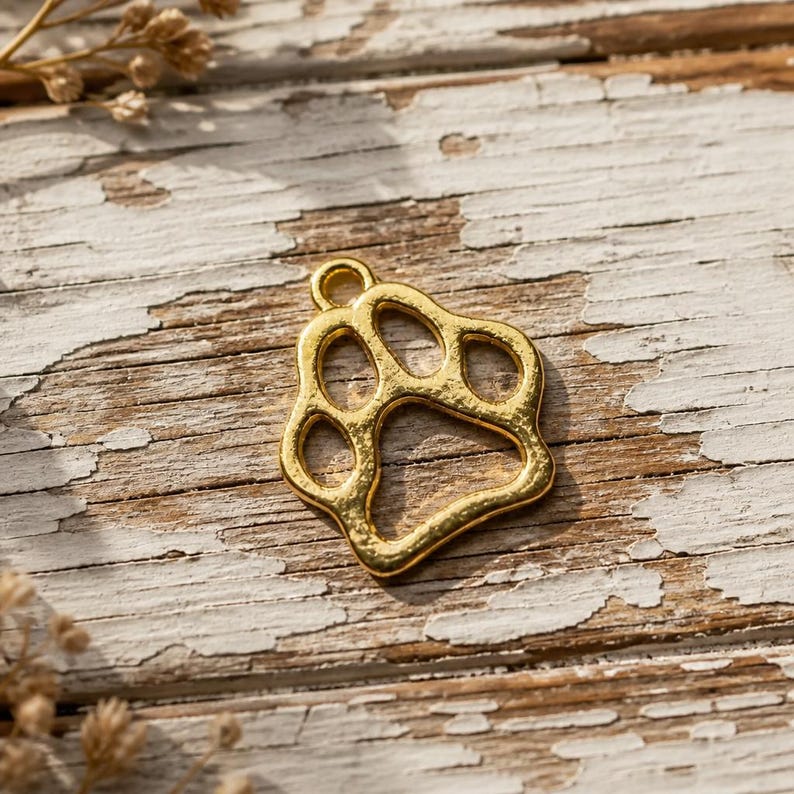 May include: A gold-toned metal charm shaped like an animal paw print. The charm features a hollow, open-work design with a small circular loop at the top for attachment. The surface of the metal has a textured, slightly uneven finish throughout.