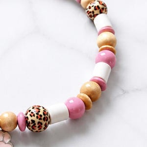 May include: A beaded necklace featuring a repeating pattern of round wooden beads in natural tan, smooth white cylindrical beads, glossy pink spherical beads, and round beads with a tan and black leopard print pattern, strung together on a cord.