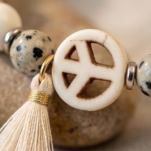 May include: A bracelet featuring round dalmatian jasper beads with black spots, silver-toned metal spacers, and a central off-white circular peace sign charm. A cream-colored thread tassel is attached to the peace sign charm with a gold-toned ring.