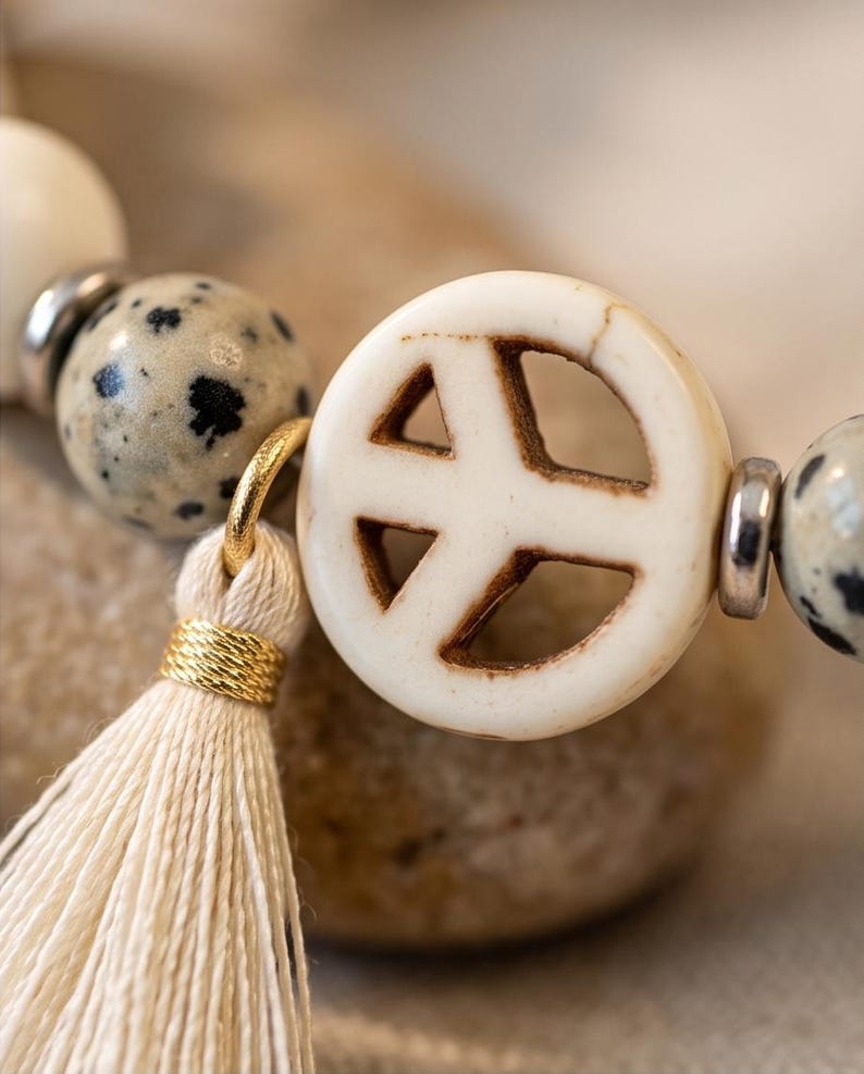 May include: A bracelet featuring round dalmatian jasper beads with black spots, silver-toned metal spacers, and a central off-white circular peace sign charm. A cream-colored thread tassel is attached to the peace sign charm with a gold-toned ring.
