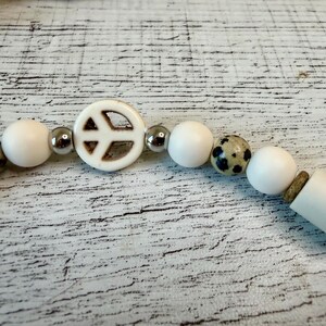 May include: A beaded bracelet featuring a central white peace sign charm, interspersed with smooth white spherical beads, metallic silver-toned spacer beads, and a single speckled tan and black bead, all strung together in a linear arrangement.
