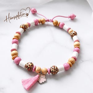 May include: A beaded bracelet featuring pink, white, and natural wood beads, along with leopard print patterned beads. The bracelet includes a pink tassel charm and a light pink paw print charm, with a pink adjustable cord and the text Hansepfoten.
