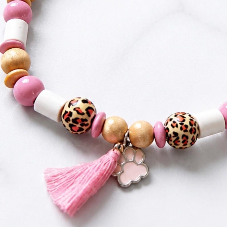 May include: A beaded bracelet featuring a sequence of pink, white, and natural wood-toned beads interspersed with leopard print spheres. The bracelet includes a dangling light pink fabric tassel and a small, light pink enamel paw print charm.
