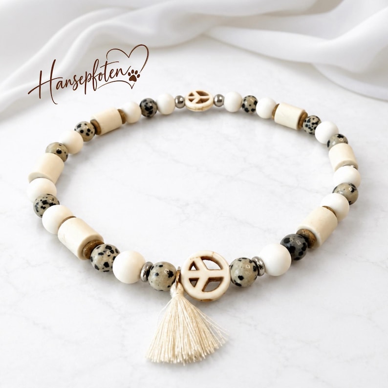May include: A beaded necklace featuring cream-colored cylindrical and spherical beads, speckled dalmatian jasper beads, and metallic spacers. The necklace includes two peace sign charms and a small cream-colored tassel. The text Hansepfoten is visible.