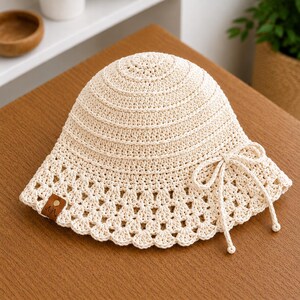 May include: A cream-colored crochet bucket hat featuring a dense, circular stitch pattern on the crown and a decorative, open-work scalloped edge along the brim. A thin matching cord is tied into a bow on the side, and a small brown leather tag is attached.