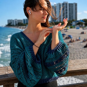 Op de afbeelding: Een teal en marineblauwe gehaakte trui met een V-hals en lange, klokmouwen. De trui heeft een open geweven ontwerp. De persoon draagt ​​witte oortelefoons en een zwarte armband. De achtergrond omvat een strand, oceaan en gebouwen.