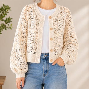 Könnte beinhalten: Eine cremefarbene, langärmelige Strickjacke mit offener Vorderseite und einem komplizierten, durchbrochenen Häkel-Blumenmuster. Das Kleidungsstück hat einen Rundhalsausschnitt, gerippte Bündchen, einen gerippten Saum und eine vertikale Knopfleiste.